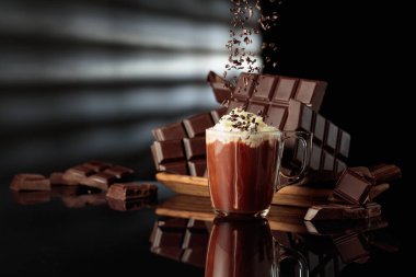 Çikolata kırıntıları serpiştirilmiş kremalı sıcak çikolata. Arka planda kırılmış acı çikolatalar. Siyah yansıtıcı arkaplan.