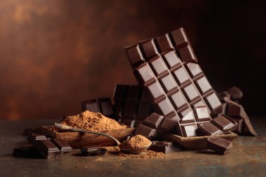 Tahta bir kase kakao tozunun yanında koyu çikolata kırıntıları. Sıcak kahverengi arka plan çikolata renginin zenginliğini vurgular. Boşluğu kopyala.