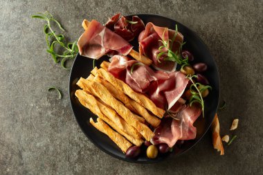 Siyah tabakta ekmek çubuğu, zeytin ve biberiyeli jambon. Taş bir masada geleneksel İtalyan atıştırmalıkları. Üst görünüm.