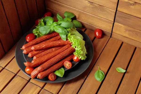 Siyah tabakta ince tütsülenmiş Polonya sosisi. Tahta bir masada fesleğen, salata ve domatesli sosisler..