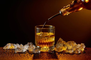 Viski şişeden cama doluyor. Viski ve buz, siyah ıslak bir masada. Sarı ışık, boşluğu kopyala.