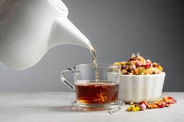 Bitki çayı cam bardağa dökülür. Kurumuş otlar ve çiçekler taş masaya saçılmış..