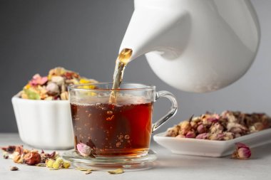 Bitki çayı cam bardağa dökülür. Kurumuş otlar ve çiçekler taş masaya saçılmış..