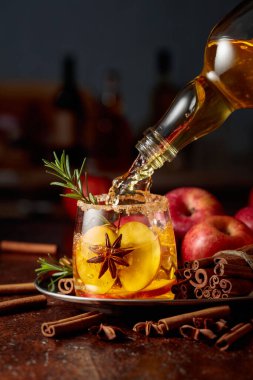 Elma şarabı bir şişeden elma dilimleri, anason ve biberiye dalıyla bir bardağa dökülür. Bardak şeker ve tarçın tozuyla süslenmiş..