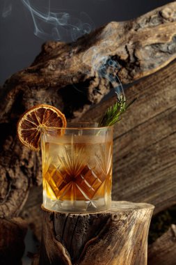 Eski usul bir kokteyl, buzlu, tahta bir kütükte. Kurutulmuş portakal dilimi ve yanan biberiye filizi ile süslenmiş soğuk, ferahlatıcı bir içecek..