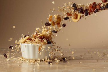 Müsli bej bir arka planda hareket halinde. Yulaf gevreği, fındık, kuru üzüm ve kuru muz dilimleri beyaz bir kaseye düşer. Boşluğu kopyala.