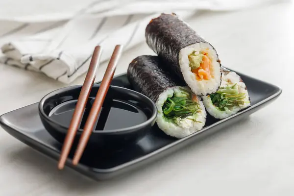 A plate filled with a variety of sushi rolls with a set of wooden chopsticks and bowl of soy sauce, placed on a simple kitchen table.