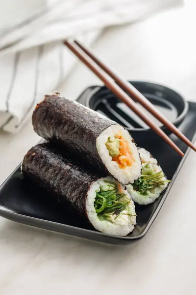A plate filled with a variety of sushi rolls with a set of wooden chopsticks and bowl of soy sauce, placed on a simple kitchen table.