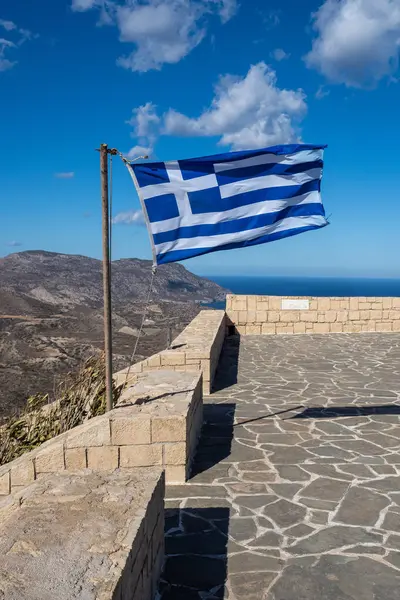 Rüzgarda dalgalanan Yunan bayrağı, dağlara ve denize tepeden bakan bir bakış açısıyla, Yunanca