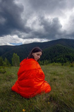 Woman tourist in orange sleeping bag  on green grass in Carpathian mountains clipart