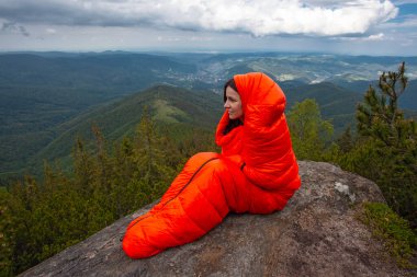 Woman tourist in orange sleeping bag on the rock in Carpathian mountains clipart