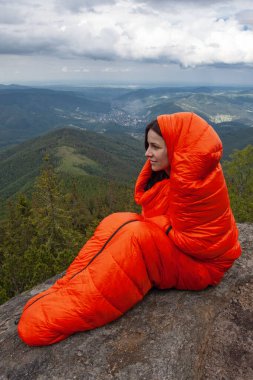 Woman tourist in orange sleeping bag on the rock in Carpathian mountains clipart