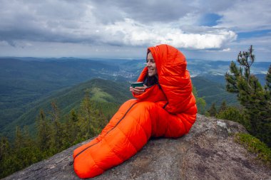 Carpathian dağlarındaki kayalıklarda turuncu uyku tulumunda kahve fincanı olan bir kadın turist.