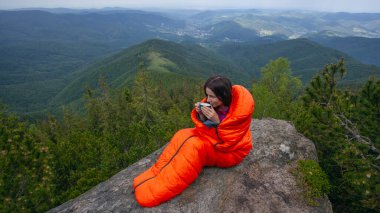 Woman tourist in orange sleeping bag with coffee cup on the rock in Carpathian mountains clipart