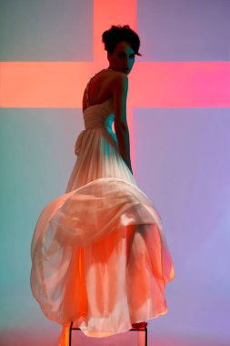 Güzel genç bayan şık bir elbise içinde fotoğraf stüdyosunda poz veriyor. Parlak renkli spot ışıklarıyla seksi bir model. Beyaz gelinlik giymiş bir kadının portresi. Sanat fotoğrafçılığı.