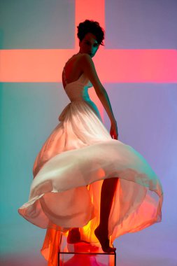 Güzel genç bayan şık bir elbise içinde fotoğraf stüdyosunda poz veriyor. Parlak renkli spot ışıklarıyla seksi bir model. Beyaz gelinlik giymiş bir kadının portresi. Sanat fotoğrafçılığı.