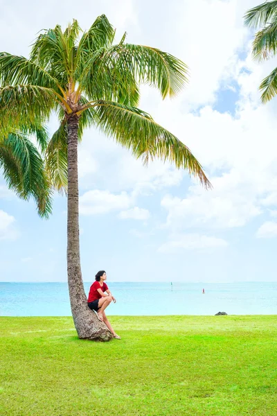穿着休闲装的年轻女子在夏威夷海旁边的草地上休息或靠着椰子或棕榈树 — 图库照片