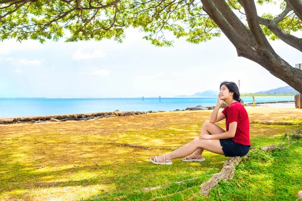 ハワイの大きなキャノピーの木の木陰で休んでいる若い女性 背景に海の地平線 — ストック写真