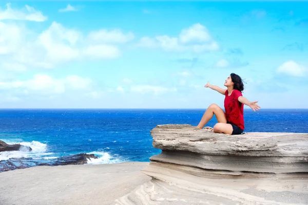 ハワイの海の大きな岩の崖に座っている若い女性は 風を楽しみながら腕を上げた — ストック写真