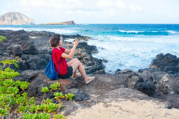 マカプーのビーチでハワイの海沿いに座っている若い女性が携帯電話で写真を撮る — ストック写真