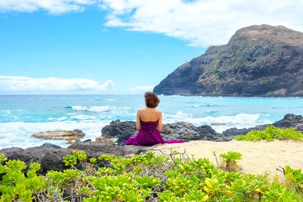 夏威夷瓦胡岛的Makapu U海滩 一名身穿紫色衣服的年轻女子坐在岩石海岸上眺望大海 图库照片