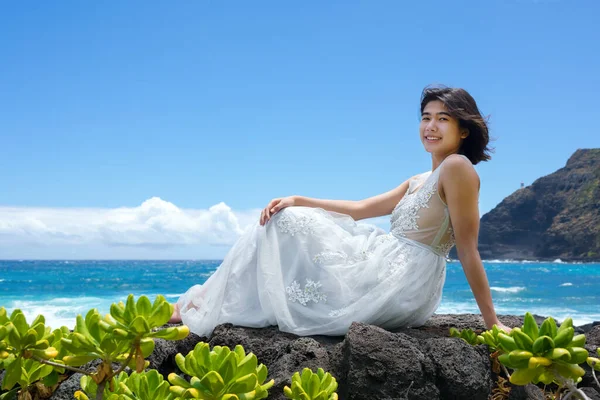 マカプーのビーチでハワイの海岸沿いの溶岩に座っている白いドレスのティーンガール ストック画像