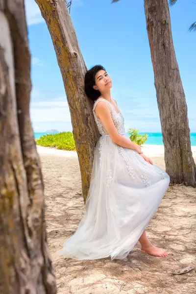 ハワイのビーチに沿って背の高い木に休む白いドレスのティーンガール 背景の青い海 ストックフォト