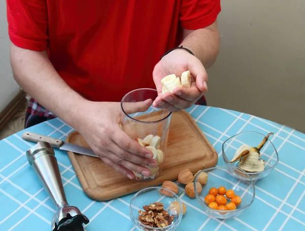 Man\'s hands preparing banana for sweet smoothie. Young cook making smoothie from banana, pieces of apples, physalis peruviana, almonds and walnuts.