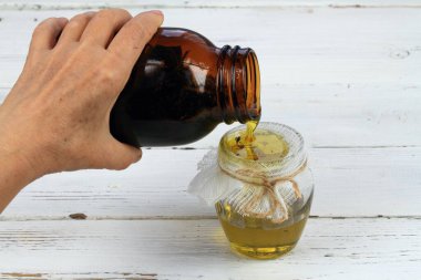 Kadının eli şişeyi tutuyor ve yağı otlarla filtreliyor. Ev yapımı bitkisel yağ beyaz masadaki cilt için iyidir..