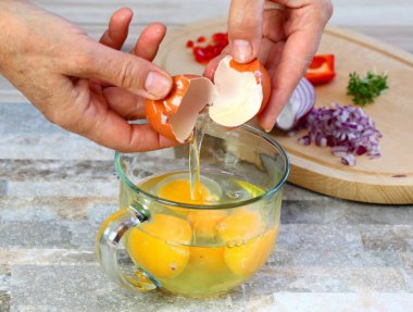 Elleri cam bardağa ve sebzeye yumurta kırıyor. Taze organik yumurtadan omlet yapmak, kırmızı soğan, kırmızı biber ve bahçe teresi kesmek..