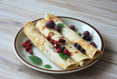 Bir tabak taze krep. Yabani çilek, mor ahududu, yabanmersini ve bitki yapraklarıyla süslenmiş krepler..