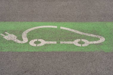 Asfalta boyanmış yeşil bir zemindeki elektrikli arabanın beyaz sembolü, EV şarj istasyonu için bir park yerini gösteriyor.