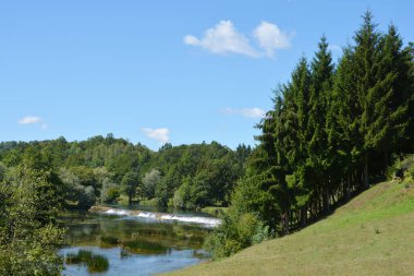 Jaskovo yakınlarındaki Dobra Nehri 'nin, mavi gökyüzünü ve beyaz bulutları yansıtan sakin suları, yumuşak bir şelale oluşturan küçük bir baraj ve kıyıları çevreleyen yemyeşil bitki örtüsü manzarası.