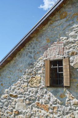 Kısmen açık pencereli, ahşap panjurlu, parlak mavi gökyüzüne karşı tek bir beyaz bulutla kurulmuş taş bir binanın ayrıntısı.