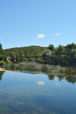 Jaskovo yakınlarındaki Dobra Nehri 'nin sakin manzarası gökyüzünü ve çevresindeki yeşillikleri yansıtan berrak sularını, küçük bir barajı ve uzaktaki eski bir köprüyü gösteriyor.