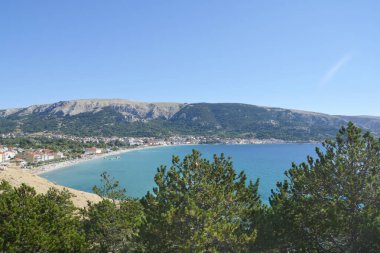 A view of Baska beach (Vela plaza) and the village of the same name, situated by the blue sea, as seen from the rocky western shore of Baska Bay in Croatia clipart