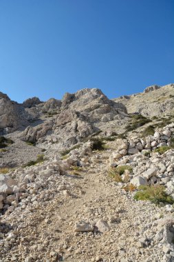 Hırvatistan 'ın Krk adasındaki Vraca geçidine (Prijevoj Vraca) uzanan kayalık tırmanışın aşağısından bir manzara, açık mavi gökyüzü altındaki engebeli taş arazinin hakimiyetinde.