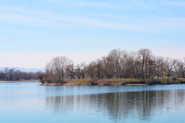 Resim Zagreb 'deki Jarun Gölü' ndeki Üniversite Adayı 'nı, yapraksız ağaçlar ve mavi gökyüzünün altında sakin bir su yüzeyi ile gösteriyor.