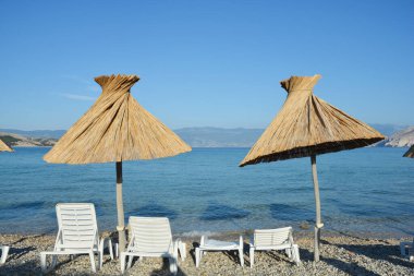 Krk Adası 'ndaki Baska' daki Vela Plaza 'nın (Büyük Plaj) bir parçası. Özgün saman şemsiyeleri ve şezlonglarıyla arka planda deniz ve dağlara bakan