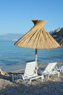 Krk Adası 'ndaki Baska' daki Vela Plaza 'nın (Büyük Plaj) bir parçası. Özgün saman şemsiyeleri ve şezlonglarıyla arka planda deniz ve dağlara bakan