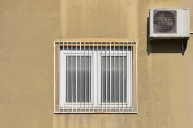 Yıpranmış bej bir bina. Kare penceresi beyaz metal parmaklıklar ve öğlen güneşinin altında bitişik bir havalandırma ünitesi tarafından korunuyor.