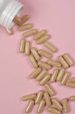 Beyaz plastik bir şişeden açık pembe arka plana bej kapsüllerin döküldüğü tepeden aşağı bir çekim.