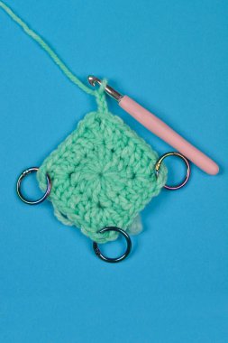 Kısmen tamamlanmış, nane yeşili iplikle örgü örme projesinin yukarıdan aşağı görüntüsü. Mavi arka planda pembe örgü kancası ve metal dikiş izleri var.