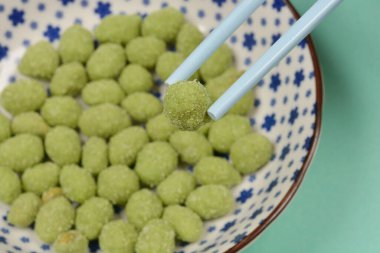 Dekoratif bir kasede wasabi kaplı fıstıklardan oluşan yakın plan bir fotoğraf. Mavi yemek çubukları arasında bir fıstık var.