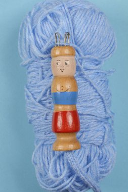 A wooden knitting doll with metal hooks, placed on a skein of blue wool, displayed against a blue background clipart