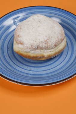 Üzerinde pudra şekeri serpiştirilmiş tek bir çöreğin, dairesel desenli mavi bir tabağa yerleştirilmiş, turuncu bir arkaplan üzerine serpilmiş,