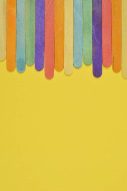 Katı sarı arkaplanın üst kenarı boyunca sıralanmış çeşitli renkli el sanatları çubuklarının yukarıdan aşağı görüntüsü canlı bir sınır oluşturur.