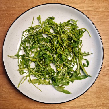 Taze roka yapraklarıyla dolu beyaz bir tabağın yukarıdan aşağı görüntüsü, ahşap bir masanın üzerine yerleştirilmiş, sadeliği içinde yeşil yapraklı sebzeleri gösteriyor.