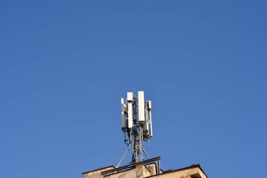 Çoklu antenleri olan bir telekomünikasyon kulesi, yıpranmış ön cephesi olan bir binanın tepesinde açık mavi bir gökyüzüne karşı duruyor.