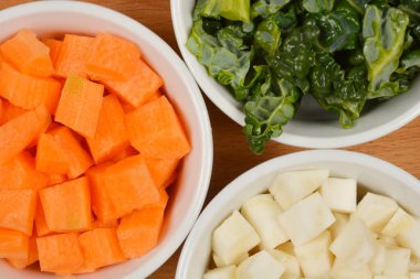 Yakın plan, tepeden tırnağa doğranmış sebzelerle dolu üç beyaz kase - ahşap bir yüzeye yerleştirilmiş savoy lahana, havuç ve kereviz kökü